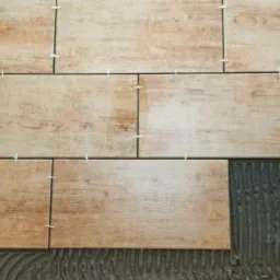 Układanie płytek imitujących drewno na ścianie, widoczne krzyżyki dystansowe i nałożony klej.