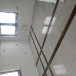 Wnętrze w trakcie prac wykończeniowych: białe płytki podłogowe z dekoracyjnymi brązowymi listwami, ściany częściowo pokryte szpachlą, widoczne okna z folią ochronną.