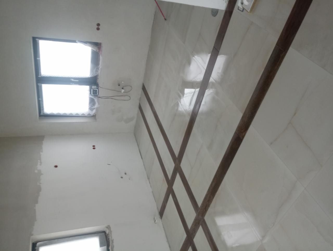 Wnętrze w trakcie prac wykończeniowych: białe płytki podłogowe z dekoracyjnymi brązowymi listwami, ściany częściowo pokryte szpachlą, widoczne okna z folią ochronną.