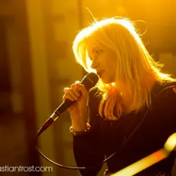 Wokalistka z blond włosami śpiewa do mikrofonu na scenie w ciepłym, żółtym oświetleniu, z widocznym kablem mikrofonowym i częścią zespołu w tle.