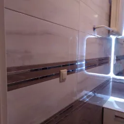 Wykończona łazienka z beżowymi płytkami imitującymi marmur, brązowymi pasami dekoracyjnymi, białym grzejnikiem drabinkowym i fragmentem białej szafki pod umywalką.