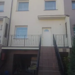 Elewacja szeregowca po renowacji, z jasną fasadą, balkonem z czarną balustradą i schodami wejściowymi, garaż w przyziemiu.