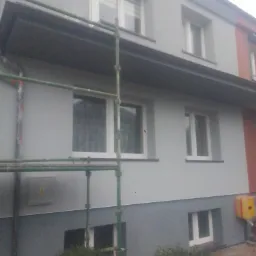 Świeżo pomalowana, dwukondygnacyjna elewacja budynku mieszkalnego w odcieniach szarości, z widocznym rusztowaniem w trakcie prac wykończeniowych.