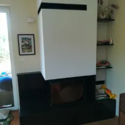 Nowoczesny kominek z czarną obudową i białym korpusem, zdobiony poziomymi czarnymi pasami, umieszczony w jasnym wnętrzu z drewnianą podłogą i półkami z dekoracjami.