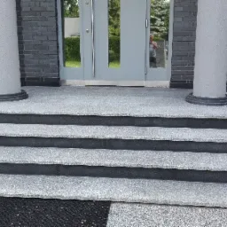 Zewnętrzne schody wejściowe z szarego kamienia z ciemnymi krawędziami, prowadzące do drzwi z kolumnami po bokach, widok z dołu.