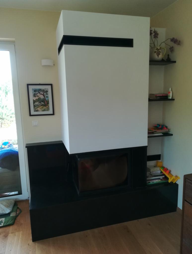 Nowoczesny kominek z czarną obudową i białym korpusem, zdobiony poziomymi czarnymi pasami, umieszczony w jasnym wnętrzu z drewnianą podłogą i półkami z dekoracjami.