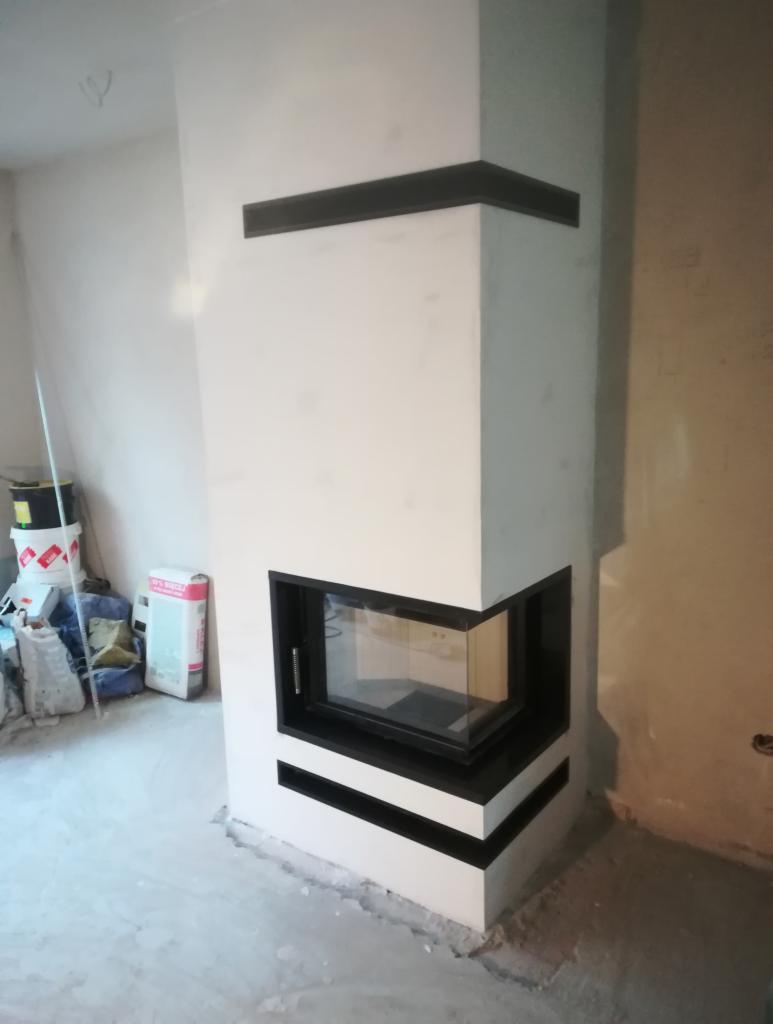Narożny kominek w trakcie wykończenia, biała obudowa z czarnymi akcentami, widoczne materiały budowlane w tle.