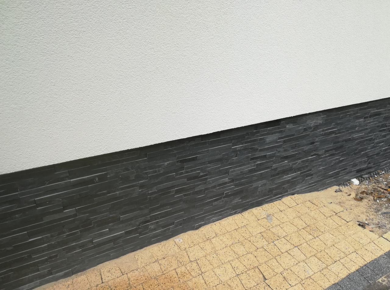 Elewacja budynku z szarego kamienia łupanego ułożonego poziomo, przylegająca do białego, teksturowanego tynku, z fragmentem chodnika z kostki brukowej w kolorze piaskowym.