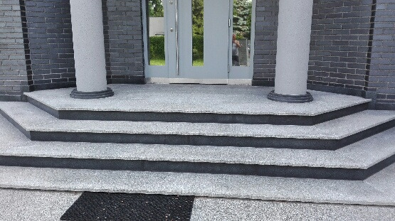Zewnętrzne schody wejściowe z szarego kamienia z ciemnymi krawędziami, prowadzące do drzwi z kolumnami po bokach, widok z dołu.