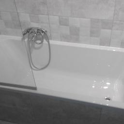 Teamlux - Biała wanna z zamontowanym szklanym parawanem nawannowym i chromowaną baterią, obudowana szarymi płytkami w łazience.