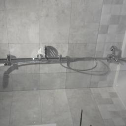 Teamlux - Szklana kabina prysznicowa z chromowaną armaturą, zamontowana w łazience we Wrocławiu wyłożonej szarymi płytkami. Widoczny panel prysznicowy z dwoma słuchawkami i baterią.