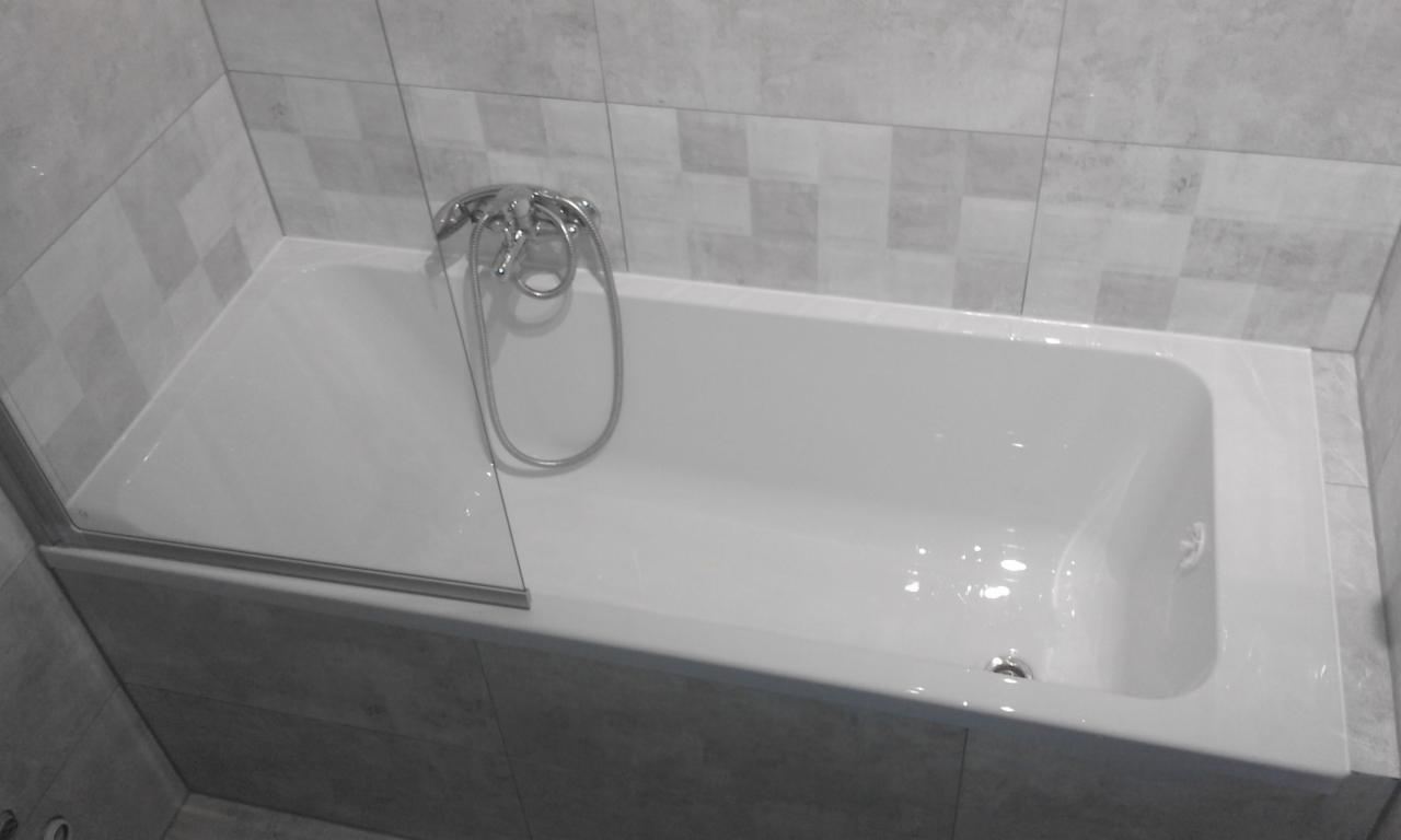 Biała wanna z zamontowanym szklanym parawanem nawannowym i chromowaną baterią, obudowana szarymi płytkami w łazience.