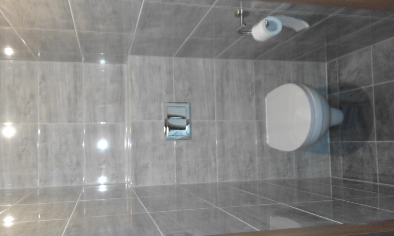 Nowoczesna toaleta wisząca w łazience wykończonej szarymi, marmurkowymi płytkami, z chromowanym przyciskiem spłukiwania i uchwytem na papier toaletowy.