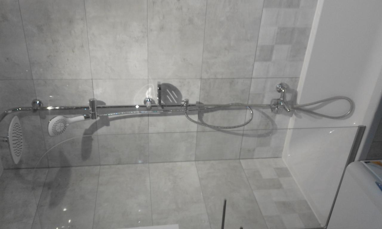 Szklana kabina prysznicowa z chromowaną armaturą, zamontowana w łazience we Wrocławiu wyłożonej szarymi płytkami. Widoczny panel prysznicowy z dwoma słuchawkami i baterią.