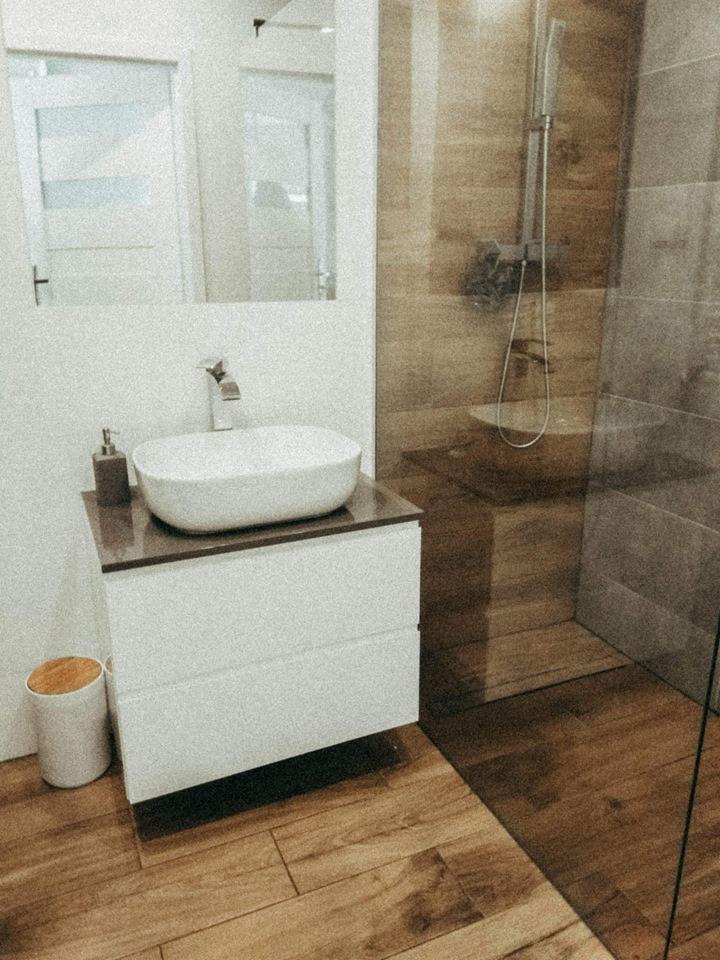 Nowoczesna łazienka z drewnianą podłogą i kabiną prysznicową typu walk-in, biała umywalka nablatowa na szafce z szufladami, widoczny dozownik mydła.