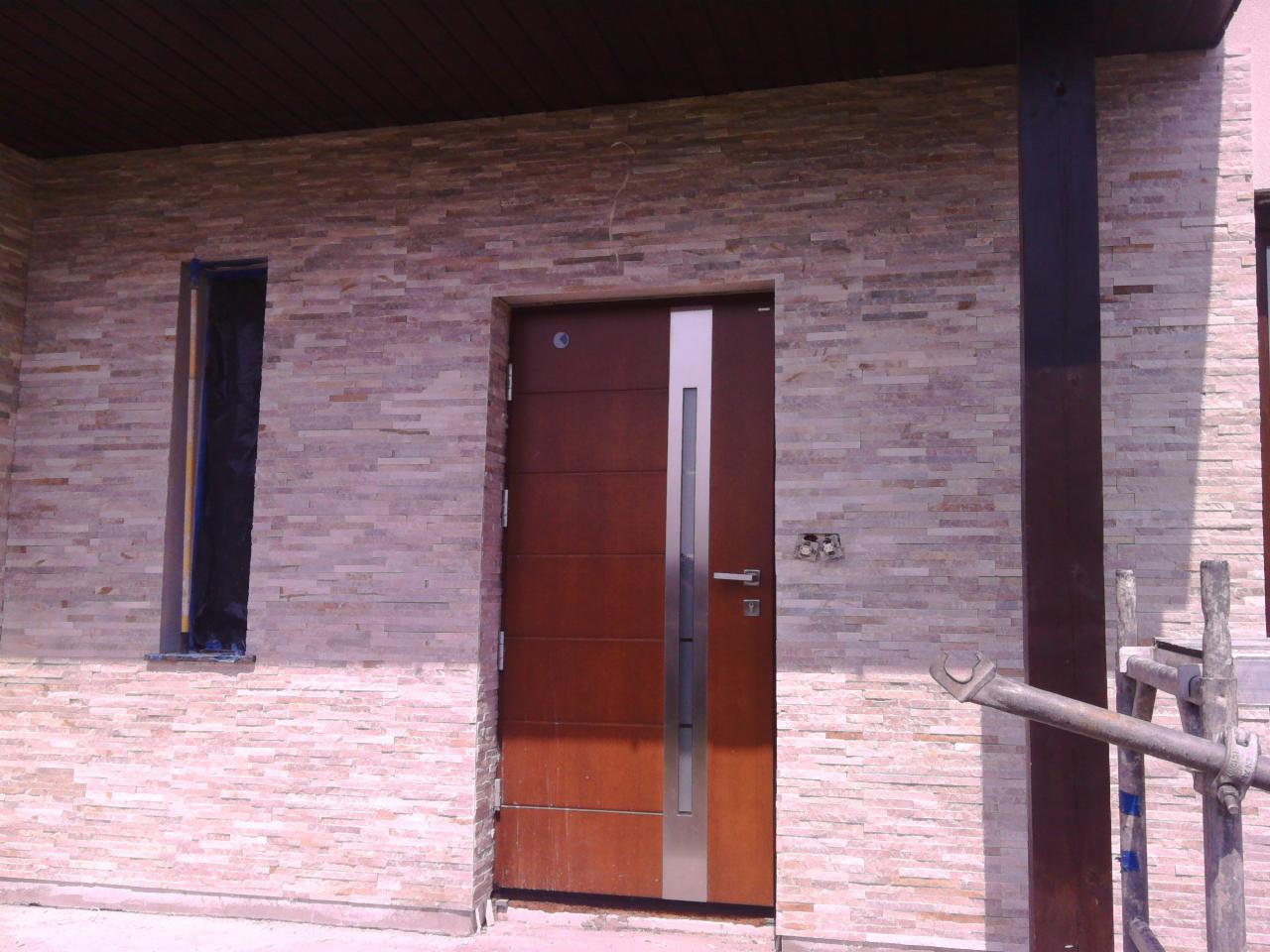 Fasada budynku wykończona kamieniem dekoracyjnym, z nowoczesnymi drzwiami wejściowymi z elementami stali nierdzewnej i oknem zabezpieczonym folią ochronną podczas prac wykończeniowych.