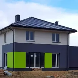 Dwukondygnacyjny dom z dachem z ciemnej dachówki, pomalowany na biało i szaro, z jaskrawozielonymi elementami elewacji na parterze, widoczne palety i cegły na ziemi.