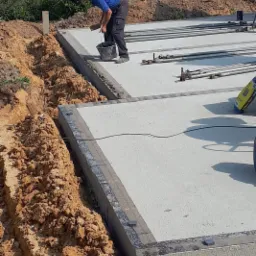 Pracownik w niebieskiej koszulce i czapce wyrównuje zaprawę na krawędzi wylewki fundamentowej, widoczny wykop, zbrojenie i worek z zaprawą.