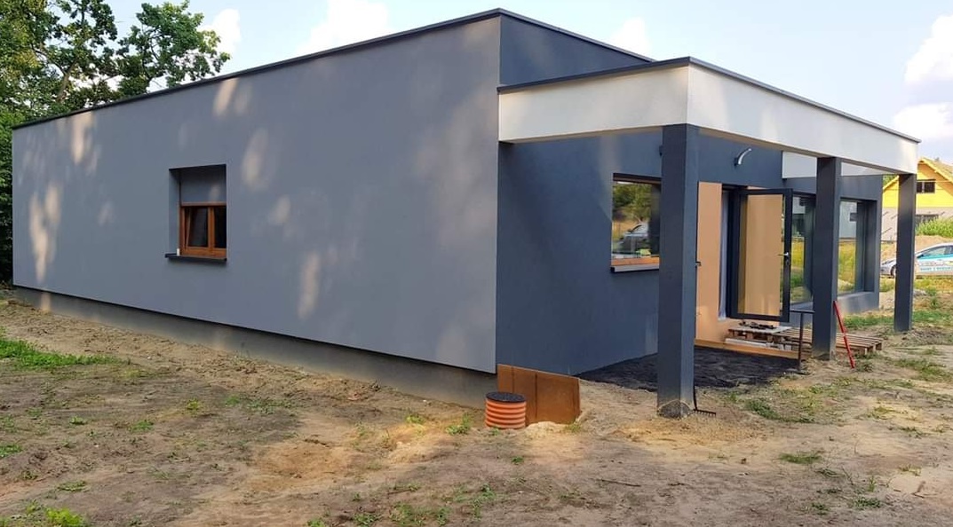 Nowoczesny, parterowy budynek z szarą elewacją, drewnianą stolarką okienną i zadaszonym wejściem, otoczony piaszczystym terenem z widocznymi elementami instalacji zewnętrznej.