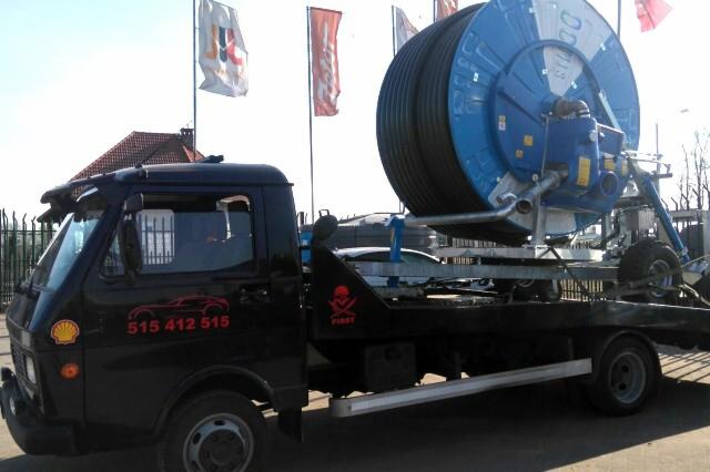 Czarna laweta z dużą niebieską rolą nawadniającą na platformie, widoczne logo Shell i numer telefonu na kabinie, tło z flagami.