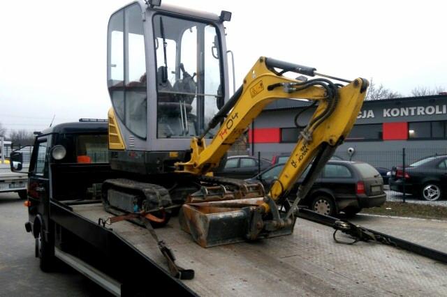 Żółto-szara mini koparka Wacker Neuson 1404, zabezpieczona pasami, przewożona na czarnej platformie pojazdu transportowego; w tle budynek z napisem 'CJA KONTROLI'.