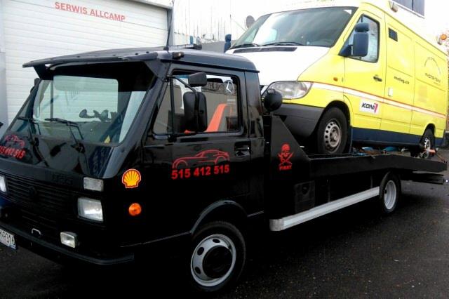 Czarna laweta z czerwonymi elementami graficznymi (samochód, numer telefonu, czaszka) przewozi biało-żółty samochód dostawczy na tle szarego budynku z napisem SERWIS ALLCAMP.