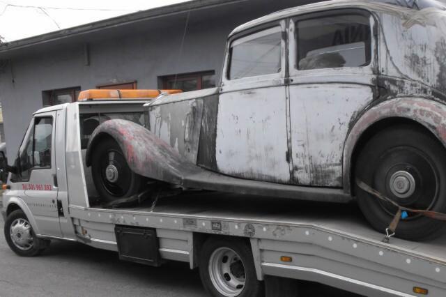 Laweta przewozi zniszczony, zabytkowy samochód osobowy. Auto jest zabezpieczone pasami. Widoczny budynek w tle.