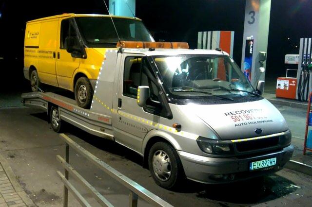 Żółty dostawczy van przewożony na srebrnej platformie pojazdu pomocy drogowej na stacji benzynowej nocą, widoczne dystrybutory paliwa i oświetlenie stacji.