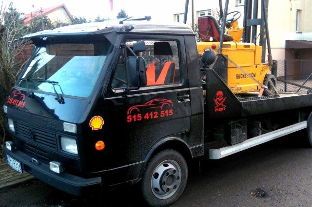 Czarna laweta z żółtym wózkiem widłowym na platformie, widoczne logo firmy z grafiką samochodu i numerem telefonu, dodatkowo naklejka z czaszką i napisem PIRAT.