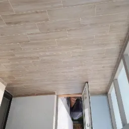 Beżowe płytki imitujące drewno ułożone na podłodze balkonu, widok z góry, z widoczną białą stolarką okienną i otwartymi drzwiami balkonowymi.