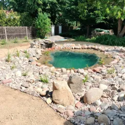 Oczko wodne z kaskadą, obłożone kamieniami różnej wielkości, otoczone świeżo posadzoną zielenią, z widoczną butelką wody przy brzegu.