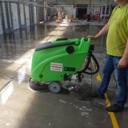 Manufaktura Czystości - Pracownik w zielonej koszulce obsługuje zieloną maszynę do mycia posadzek w hali przemysłowej, widoczne mokre ślady po czyszczeniu i żółte pasy na podłodze.