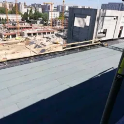 Widok z góry na płaski dach pokryty szarą membraną hydroizolacyjną, w tle plac budowy z dźwigami i budynkami w trakcie wznoszenia, fragment rusztowania w prawym dolnym rogu.