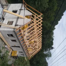 Surowy stan domu z białych bloczków z drewnianą konstrukcją dachu w trakcie budowy, widok z góry z elementami zieleni i samochodem dostawczym w tle.