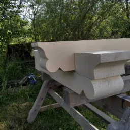 Element drewniany z dekoracyjnymi wycięciami, prawdopodobnie fragment konstrukcji dachowej, leżący na drewnianym koźle w otoczeniu zieleni.