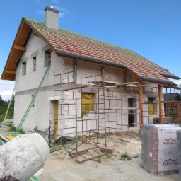 Niedokończony dom z pustaków, z częściowo położoną czerwoną dachówką i przygotowaną konstrukcją dachu, widoczne rusztowania, betonowa mieszarka i paleta z dachówką Braas