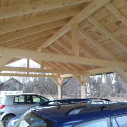 Drewniana konstrukcja zadaszenia nad dwoma samochodami, widoczna więźba dachowa i poszycie z desek, w tle drzewa bez liści.