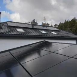 Panele fotowoltaiczne zamontowane na dachu domu z dachówką i oknami dachowymi, widok z bliska na część instalacji, pochmurne niebo w tle.