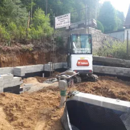 Wykop pod fundamenty z częściowo wzniesionymi ścianami z bloczk&oacute;w betonowych, zabezpieczonych czarną folią hydroizolacyjną, ubijarka wibracyjna i minikoparka Bobcat na placu budowy, w tle zieleń...
