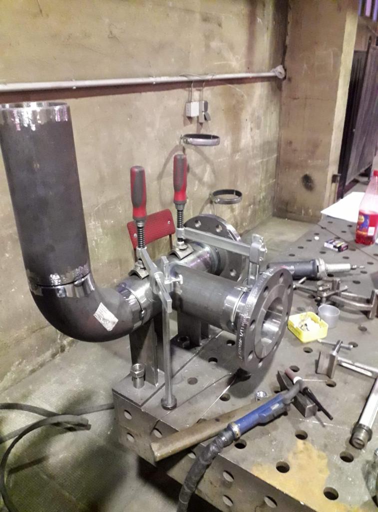 Spawanie rur z kołnierzami na stole spawalniczym z użyciem uchwytów zaciskowych, widoczne narzędzia i akcesoria spawalnicze.