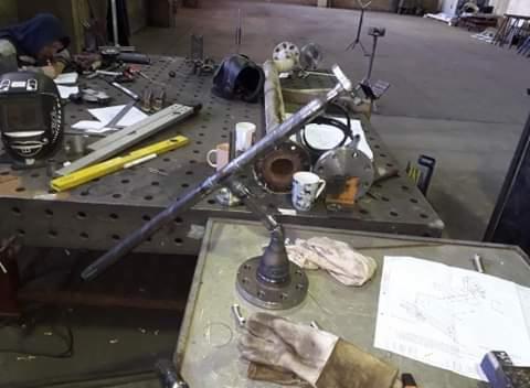 Warsztatowa przestrzeń robocza z metalowymi elementami konstrukcyjnymi w trakcie obróbki, widoczna maska spawalnicza, poziomica, kubki oraz rysunek techniczny na stole.