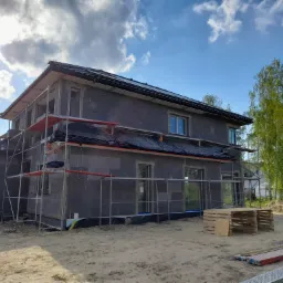Dom jednorodzinny w Radomiu w trakcie ocieplania, widoczne rusztowania, szary styropian na ścianach, czarna folia na dachu oraz elementy elewacyjne przygotowane do montażu.