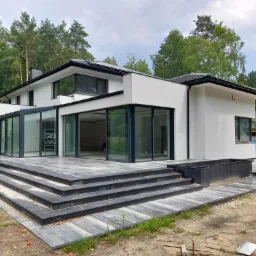 Nowoczesny, dwukondygnacyjny dom z białym wykończeniem elewacji, dużymi przesuwnymi oknami i schodami prowadzącymi na taras, otoczony zielenią drzew.