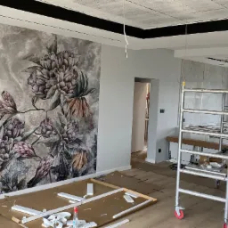 Wnętrze w trakcie malowania: ściana z tapetą w kwiaty, rozłożone listwy przypodłogowe, rusztowanie malarskie oraz zabezpieczona podłoga.