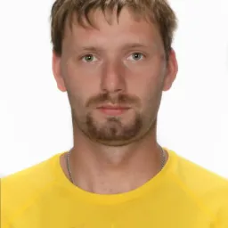 Portret mężczyzny z krótkimi, jasnymi włosami i zarostem, ubranego w żółtą koszulkę sportową z nadrukiem. Białe tło, neutralny wyraz twarzy.