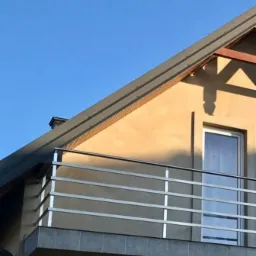 Balkon z nowoczesną, poziomą balustradą ze stali na tle jasnego budynku z ciemnym dachem i błękitnym niebem.