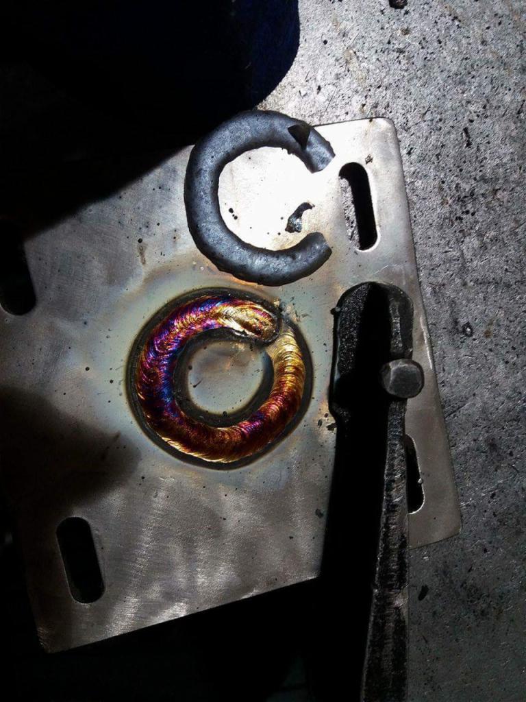 Metalowa płytka z otworem i przyspawanym pierścieniem, widoczne tęczowe zabarwienia spoiny, obok leży fragment metalu w kształcie litery C i narzędzie zaciskowe.