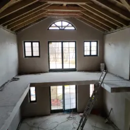 Wnętrze domu w stanie surowym zamkniętym po tynkowaniu maszynowym, widoczne drewniane elementy więźby dachowej, okna z czarnymi ramami, schody i rusztowanie.