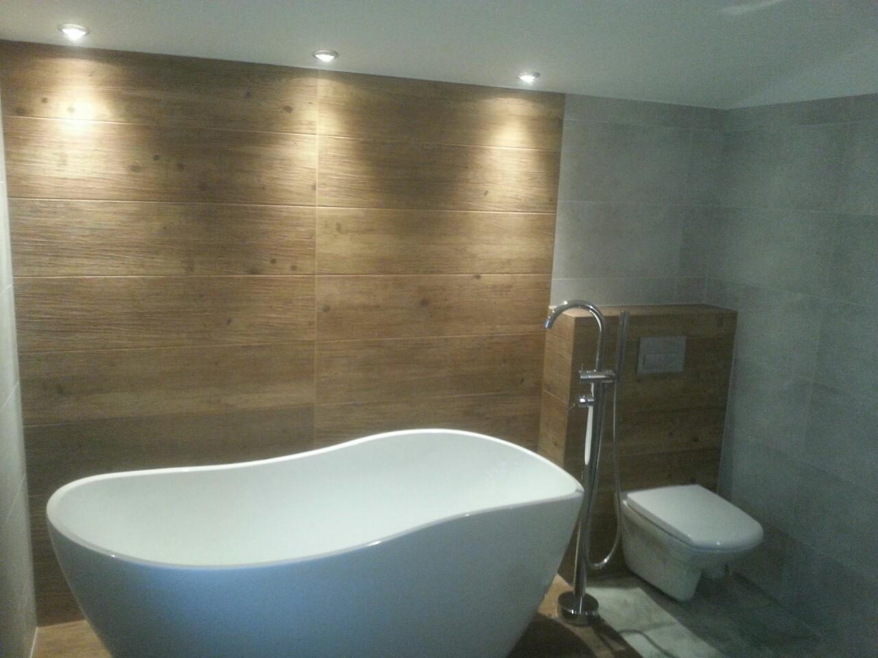 Nowoczesna łazienka z wolnostojącą białą wanną, baterią podłogową i drewnopodobną okładziną ściany z podświetleniem punktowym.