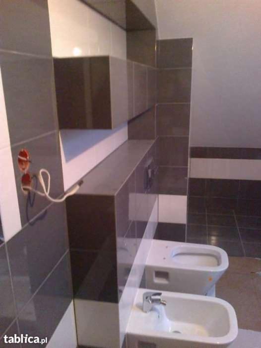 Ukończona łazienka z szaro-białymi płytkami, widoczna toaleta i bidet, niedokończone gniazdko elektryczne z wystającymi kablami sugerujące, że prace jeszcze trwają.